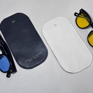 May include: Two pairs of sunglasses with black frames and colored lenses, one with blue and the other with yellow lenses, are displayed with two leather eyeglass cases. One case is navy blue, the other is white.