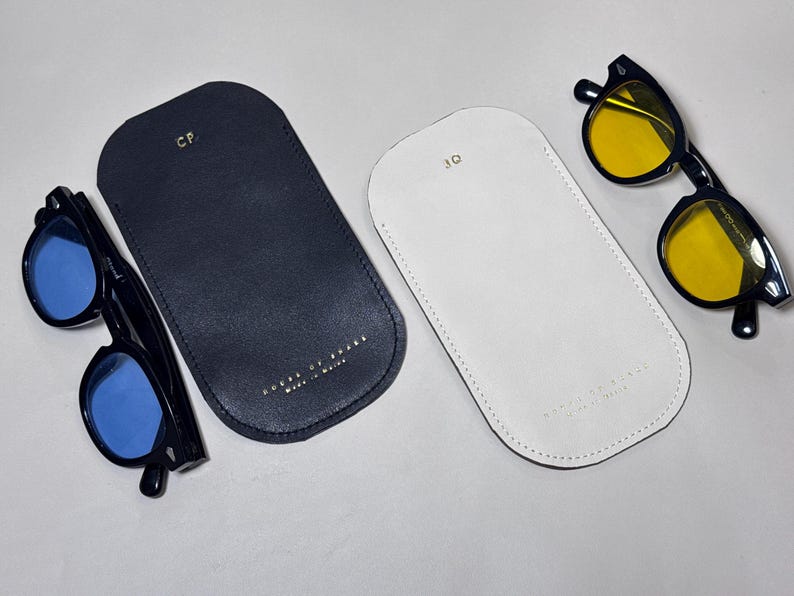 May include: Two pairs of sunglasses with black frames and colored lenses, one with blue and the other with yellow lenses, are displayed with two leather eyeglass cases. One case is navy blue, the other is white.