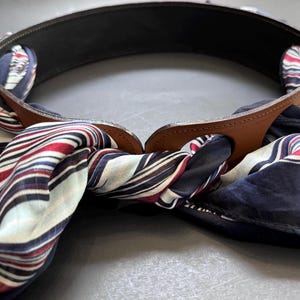 Puede incluir: Un cinturón de cuero negro con un acento de cuero marrón y una bufanda a rayas en rojo, blanco y azul marino. La bufanda está pasada por el cinturón, creando un accesorio elegante.