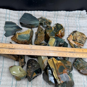 May include: A collection of raw, multi-coloured jasper stones in shades of green, brown, yellow, and grey, displayed with a wooden ruler showing measurements in centimetres, with the stones ranging from approximately 2 to 8 cm in size.