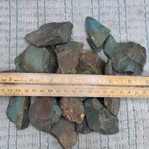 May include: A collection of raw, angular stones in shades of sage green, brown, and gray, positioned beneath a wooden ruler that measures approximately 7 inches across the width of the stone cluster, showing varied textures and natural mineral layers.