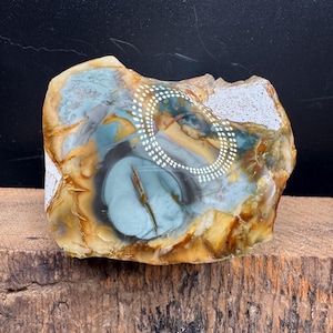 May include: A polished stone with a unique pattern. The stone features a mix of colors, including tan, blue, and brown. A white dotted design is visible on the surface. The stone is displayed on a wooden surface.