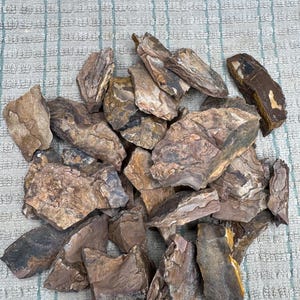 May include: A collection of raw, jagged stones in various shades of brown, tan, and grey. The rocks feature natural, uneven textures, sharp edges, and layered mineral patterns with earthy tones, displaying a rough, unpolished, and organic appearance.
