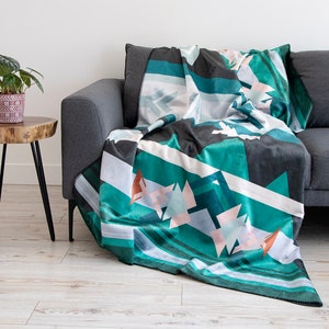 Peut inclure: Une couverture à motifs géométriques dans les tons de turquoise, gris, blanc et corail est drapée sur un canapé gris. La couverture présente un motif répétitif de formes angulaires et de rayures. Une petite plante en pot est posée sur une table d'appoint en bois.