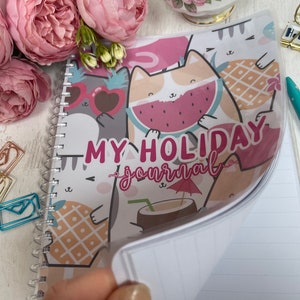 Puede incluir: Un cuaderno de espiral rosa y blanco con una portada colorida que presenta gatos de dibujos animados, piñas, sandía y una sombrilla de playa. El texto de la portada dice "My Holiday Journal".