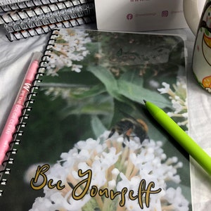 Op de afbeelding: Een spiraalgebonden notitieboek met een doorzichtige plastic hoes met een close-up foto van witte bloemen en een bij. De tekst "Bee Yourself" staat in gouden letters op de hoes.