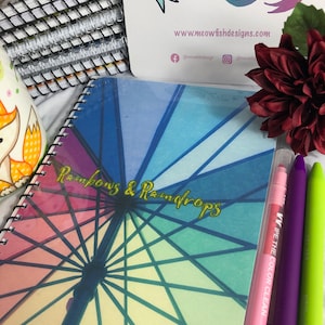 Könnte beinhalten: Ein Spiralnotizbuch mit einem Regenbogen-Schirm-Design und den goldenen Worten "Rainbows & Raindrops". Das Bild zeigt auch einen rosa, lila und grünen Stift, eine Visitenkarte mit einem Katzen-Meerjungfrau-Logo und eine dekorative Blume.