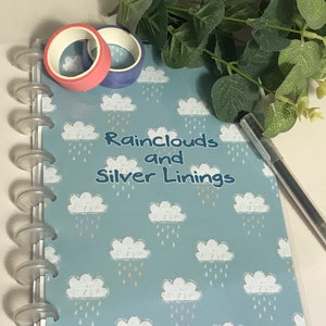 Może przedstawiać: Niebieski notes z wzorem białych chmur i tekstem "Rainclouds and Silver Linings". Notes ma przezroczystą plastikową okładkę i srebrny segregator z pierścieniami. Na notesie leżą dwa rolki taśmy washi, jedna różowa i jedna niebieska.