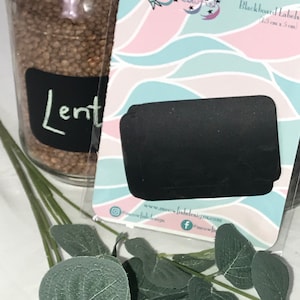 Pode incluir: Um frasco de lentilhas com um rótulo de quadro negro preto que diz "Lentils" em giz branco. Um pacote de etiquetas de quadro negro, 15 cm x 5 cm, está em primeiro plano. O pacote tem um design pastel com um logotipo de gato.