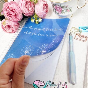 Puede incluir: Una libreta espiral azul con una cita que dice "El momento perfecto para hacer lo que amas es ahora." La libreta tiene una cubierta de plástico transparente con un diseño de diente de león. La libreta está abierta en una página en blanco con papel rayado. Una ilustración de sirena es visible en la esquina inferior derecha de la página.