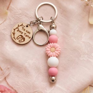 May include: A silver keychain with a round wooden charm engraved with the text "Tu vas &ecirc;tre Tata" and baby footprints. The keychain features pink and white beads, a pink daisy, and a silver clasp.