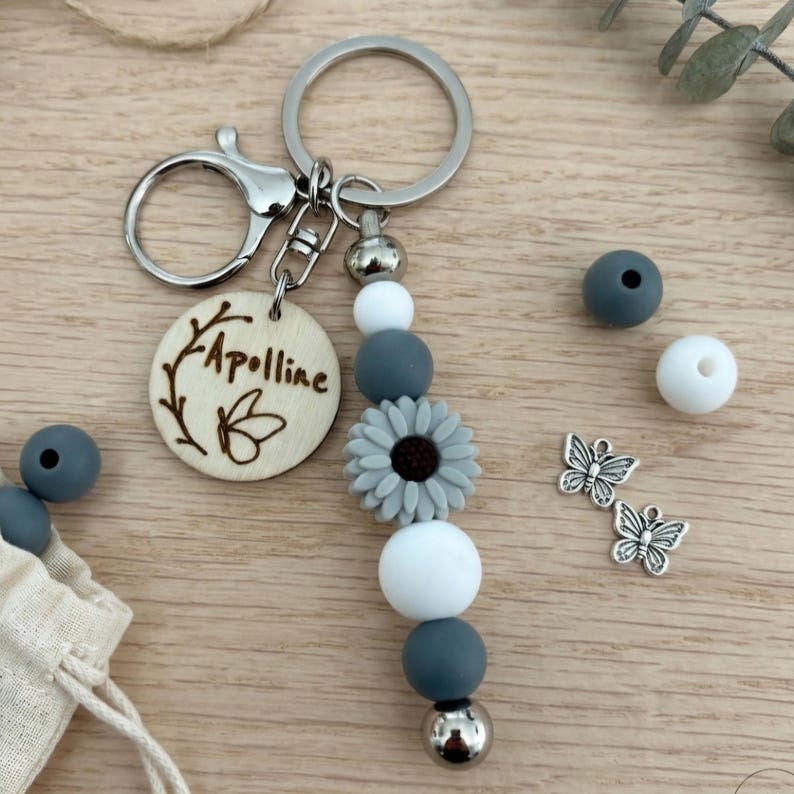 May include: A handmade keychain with a wooden charm engraved with the name "Apolline" and a butterfly design. The keychain features gray, white, and silver beads, a gray flower, and silver butterfly charms. The keychain is attached to a silver keyring.