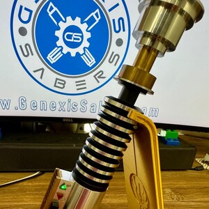May include: A gold lightsaber prop with a black and silver handle, a green light, and a gold stand featuring a Mandalorian symbol. The stand is on a wooden surface. The lightsaber is on display and is part of a collection of Star Wars memorabilia.