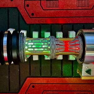 May include: A close-up of a lightsaber hilt with a green and red light saber blade. The hilt is made of metal and has a black and silver design. The lightsaber is on a red and black background.