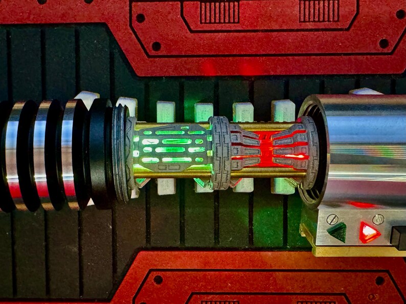 May include: A close-up of a lightsaber hilt with a green and red light saber blade. The hilt is made of metal and has a black and silver design. The lightsaber is on a red and black background.