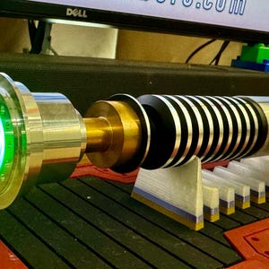 May include: A green lightsaber with a silver and gold hilt, and a black and silver blade. The lightsaber is on a white stand with a blue and gold base.