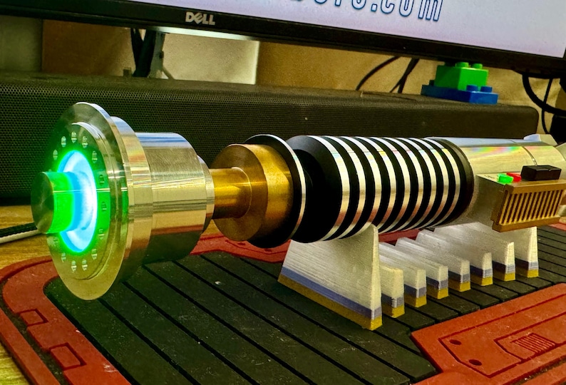 May include: A green lightsaber with a silver and gold hilt, and a black and silver blade. The lightsaber is on a white stand with a blue and gold base.