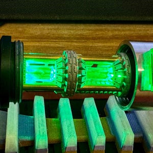 May include: A close-up of a lightsaber hilt with a green LED light. The hilt is made of metal and has a black and silver finish. The lightsaber is on a wooden surface.