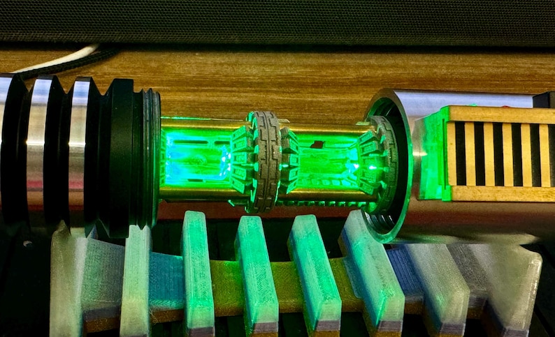 May include: A close-up of a lightsaber hilt with a green LED light. The hilt is made of metal and has a black and silver finish. The lightsaber is on a wooden surface.