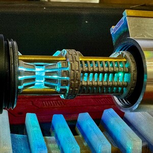 May include: A close-up of a lightsaber hilt with a blue lightsaber blade. The hilt is made of metal and has a detailed design. The lightsaber is on a blue and grey surface.