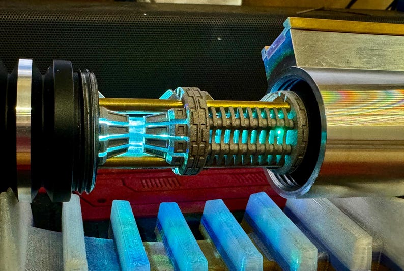 May include: A close-up of a lightsaber hilt with a blue lightsaber blade. The hilt is made of metal and has a detailed design. The lightsaber is on a blue and grey surface.