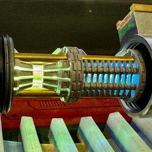 May include: A close-up of a lightsaber hilt with a gold and silver finish. The hilt has a blue light emitting from the centre. The hilt is surrounded by a black and silver casing.
