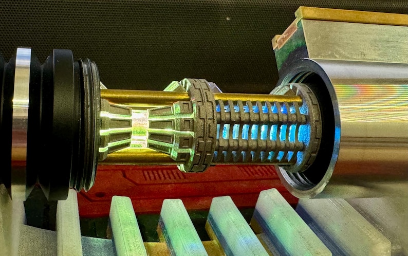 May include: A close-up of a lightsaber hilt with a gold and silver finish. The hilt has a blue light emitting from the centre. The hilt is surrounded by a black and silver casing.