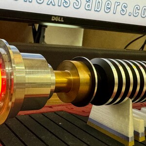 May include: A lightsaber with a black and silver hilt, a gold metal connector, and a red LED light. The lightsaber is on a stand with a wooden base.
