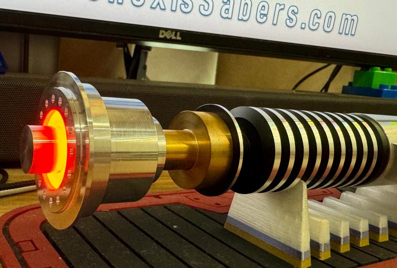 May include: A lightsaber with a black and silver hilt, a gold metal connector, and a red LED light. The lightsaber is on a stand with a wooden base.
