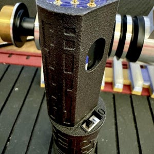May include: A black 3D printed device with a gold-coloured connector on the top and a USB connector on the bottom. The device has a textured surface and a small hole on the side.