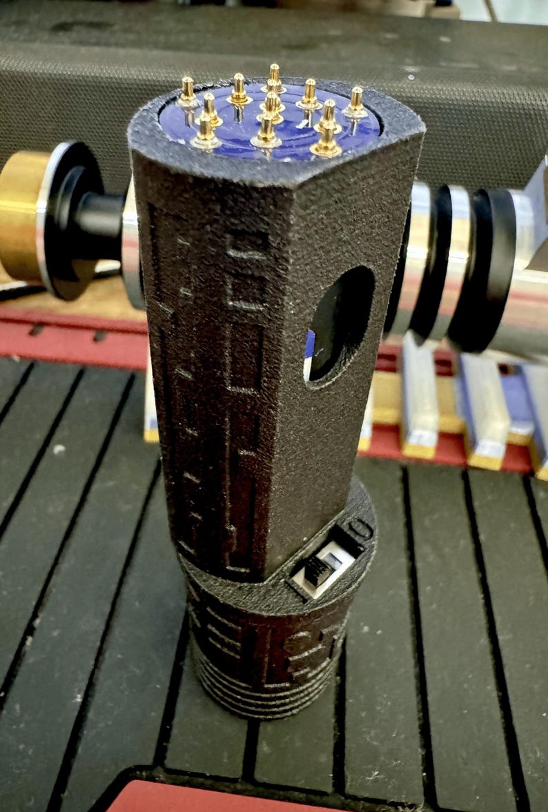 May include: A black 3D printed device with a gold-coloured connector on the top and a USB connector on the bottom. The device has a textured surface and a small hole on the side.