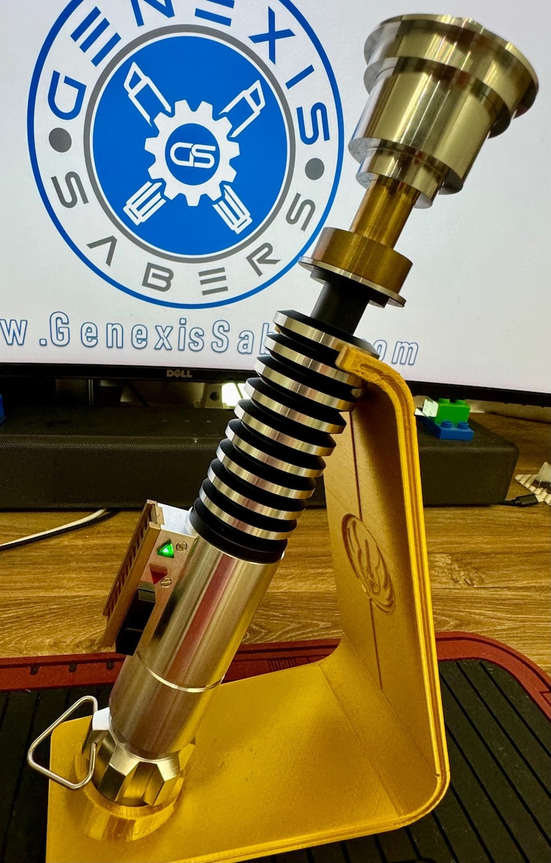 May include: A gold lightsaber prop with a black and silver handle, a green light, and a gold stand featuring a Mandalorian symbol. The stand is on a wooden surface. The lightsaber is on display and is part of a collection of Star Wars memorabilia.