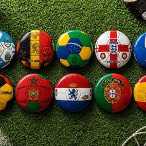Puede incluir: Una colección de balones de fútbol con banderas de diferentes países, como Argentina, Bélgica, Alemania, Marruecos, Países Bajos, Portugal y España. Los balones están colocados sobre una superficie verde, con un balón de fútbol en el fondo.
