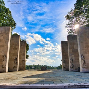 May include: A row of tall, stone pillars with sculpted figures stand in a park setting. The pillars are arranged in a symmetrical pattern, creating a walkway through the center. The sky is blue with white clouds, and the sun is shining brightly.