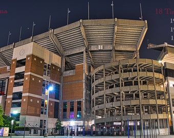 Könnte beinhalten: Eine nächtliche Außenansicht des Bryant-Denny Stadiums, der Heimat des American-Football-Teams der University of Alabama Crimson Tide. Das Stadion ist beleuchtet und der Text "University of Alabama" und "Bryant-Denny Stadium" ist oben im Bild sichtbar. Der Text "18 Time, National Champions" ist rechts im Bild sichtbar, zusammen mit einer Liste der Jahre, in denen das Team nationale Meisterschaften gewonnen hat.