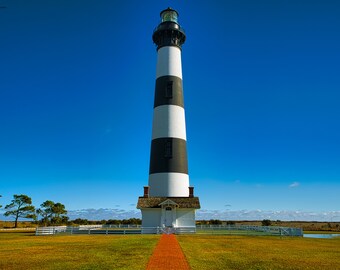 May include: A black and white striped lighthouse with a red roof stands tall against a bright blue sky. A brick path leads up to the lighthouse, which is surrounded by a white fence.