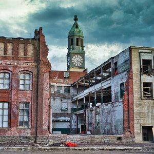 May include: A weathered brick building with a clock tower, showing signs of decay and neglect. The building has large windows and a partially collapsed roof. The sky is overcast and stormy.