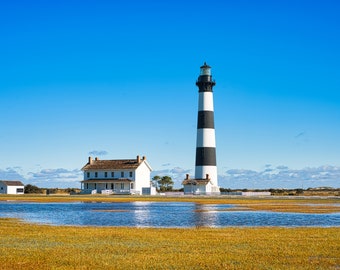 May include: A black and white striped lighthouse stands tall against a bright blue sky. A white house with a brown roof sits in front of the lighthouse. The scene is set against a backdrop of golden grass and a shallow pool of water.