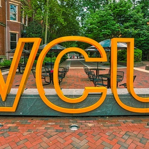 May include: A large, yellow, three-dimensional sign spelling out "VCU" on a brick walkway. The sign is in front of a brick building with trees and a courtyard behind it.