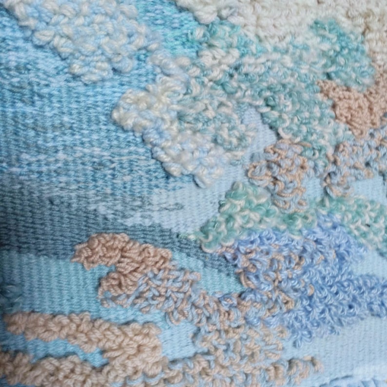 K&ouml;nnte beinhalten: Nahaufnahme einer gewebten Wandbeh&auml;nge mit einem strukturierten, abstrakten Design. Die Beh&auml;nge zeigt eine Vielzahl von Blau-, Gr&uuml;n- und Beiget&ouml;nen.