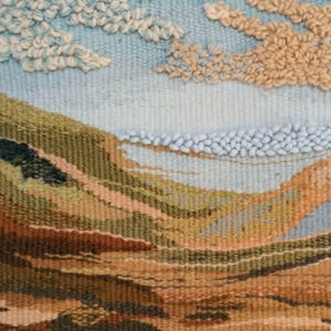 K&ouml;nnte beinhalten: Eine gewebte Wandteppich, die eine Landschaft mit blauem Himmel und flauschigen Wolken darstellt. Die Landschaft besteht aus braunen, gr&uuml;nen und orangefarbenen H&uuml;geln. Der Himmel ist hellblau mit wei&szlig;en Wolken. Die Wolken sind aus flauschigem Garn gefertigt.