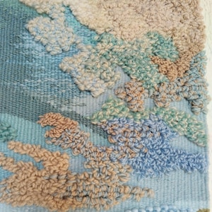 K&ouml;nnte beinhalten: Nahaufnahme einer gewebten Wandbeh&auml;nge mit einem strukturierten, abstrakten Design. Die Beh&auml;nge zeigt eine Vielzahl von Farben, darunter Blau, Gr&uuml;n und Beige.