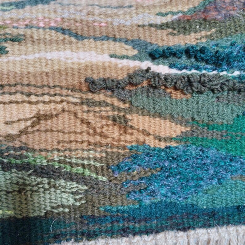 K&ouml;nnte beinhalten: Nahaufnahme einer gewebten Wandteppich mit einem Landschaftsdesign. Der Wandteppich zeigt Braunt&ouml;ne, Gr&uuml;nt&ouml;ne und Blaut&ouml;ne, die ein strukturiertes und komplexes Muster erzeugen.