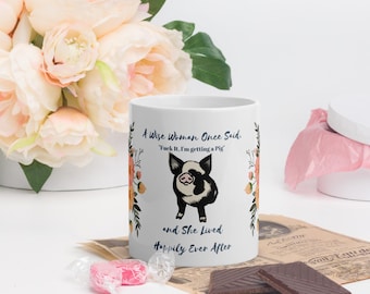 May include: A white ceramic mug featuring a black and white pig illustration centered between floral designs. The text reads: A Wise Woman Once Said, Fuck It, I'm getting a Pig, and She Lived Happily Ever After. The mug is decorated with orange flowers.