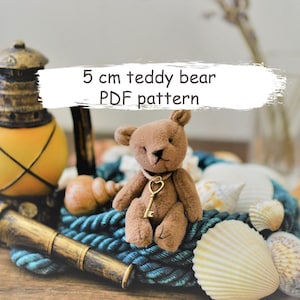 May include: A 5 cm teddy bear PDF pattern. The pattern is for a brown teddy bear with a gold key charm. The bear is sitting on a blue and green rope with seashells around it.