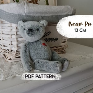 May include: A gray plush teddy bear with a red heart on its chest, sitting on a white wicker basket. The bear is 13 inches tall and has a stitched nose and mouth. The text "Bear Po 13 CM" and "PDF PATTERN" are visible in the image.