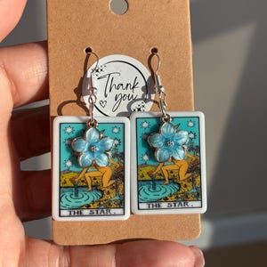 Puede incluir: Pendientes plateados con diseños rectangulares de cartas del tarot, fondo turquesa y adornos florales. Las cartas dicen "THE STAR". Los pendientes están sujetos a una tarjeta marrón con un mensaje de "Thank you".