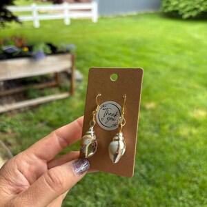 May include: A pair of seashell earrings with gold-colored hooks and accents, displayed on a brown card. The earrings have a natural, pearlescent appearance. A small sticker with the words "Thank you" is visible.