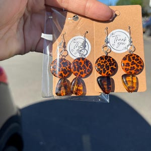 Puede incluir: Pendientes hechos a mano con un diseño de estampado de leopardo. Los pendientes tienen una parte superior circular naranja con manchas negras y una parte inferior en forma de lágrima de color ámbar. Se muestran en una tarjeta marrón con una pegatina "Thank you".