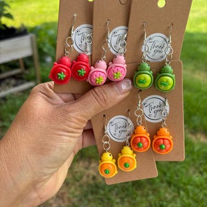 Puede incluir: Cinco pares de pendientes colgantes con dijes en forma de tortuga y caparazones con motivos de frutas en rojo, rosa, verde, naranja y amarillo. Cada par está sujeto a una tarjeta de presentación de cartón marrón con una pegatina Thank you.
